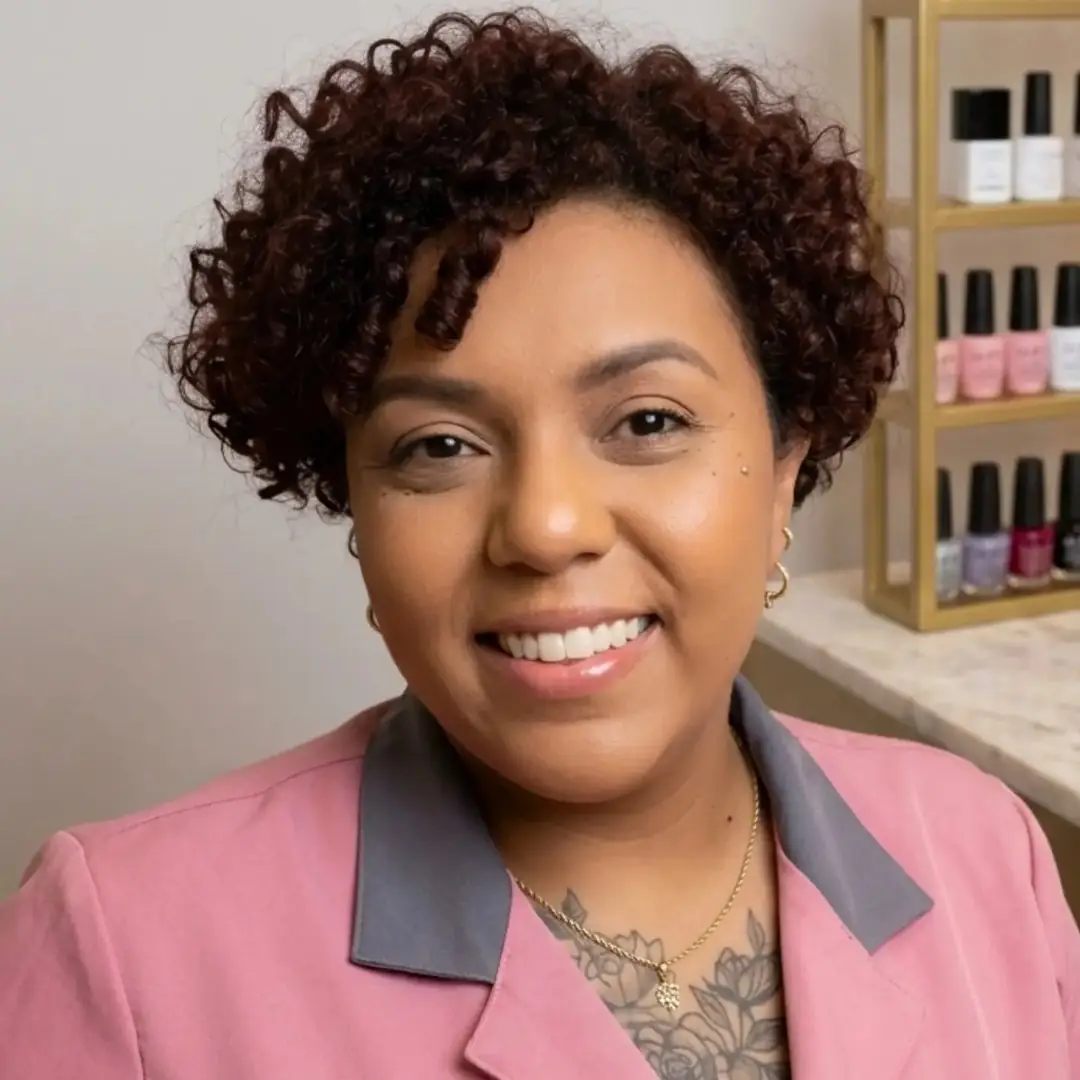Retrato de Amanda Niak sorrindo, com blazer rosa, ao lado de uma estante de esmaltes